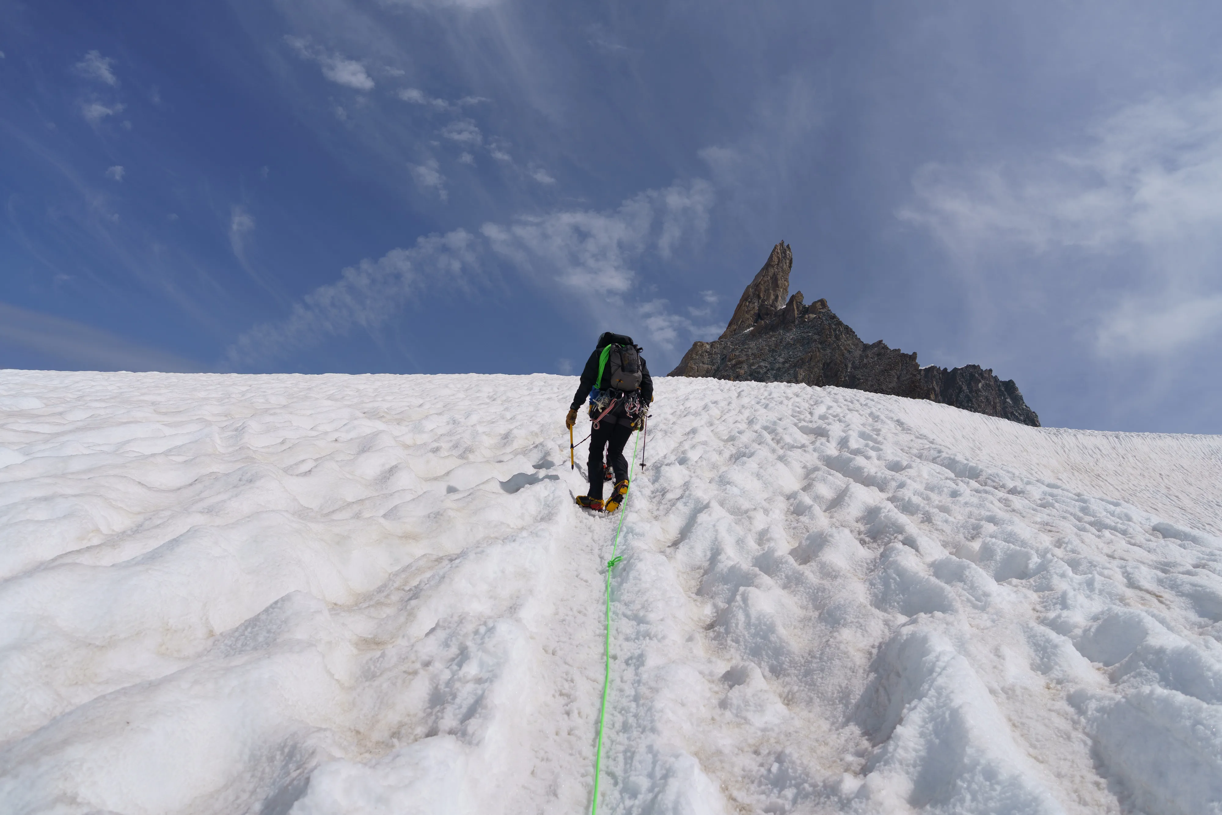 A roped climber in crampons walks up a snow slope toward the dark rock tower of the Dent du Géant; a green rope trails back down the track.